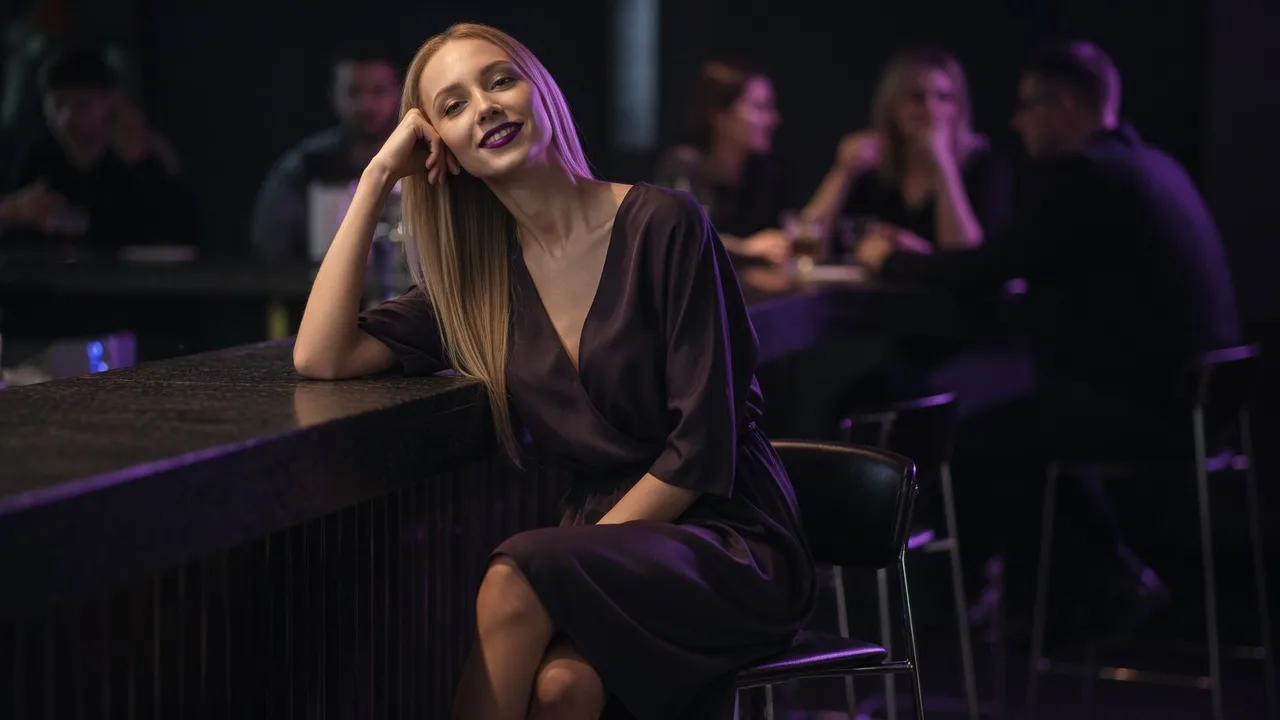A smiling transgender woman with long blonde hair, dressed in an elegant dress, sitting at a bar and looking coyly toward the observer
